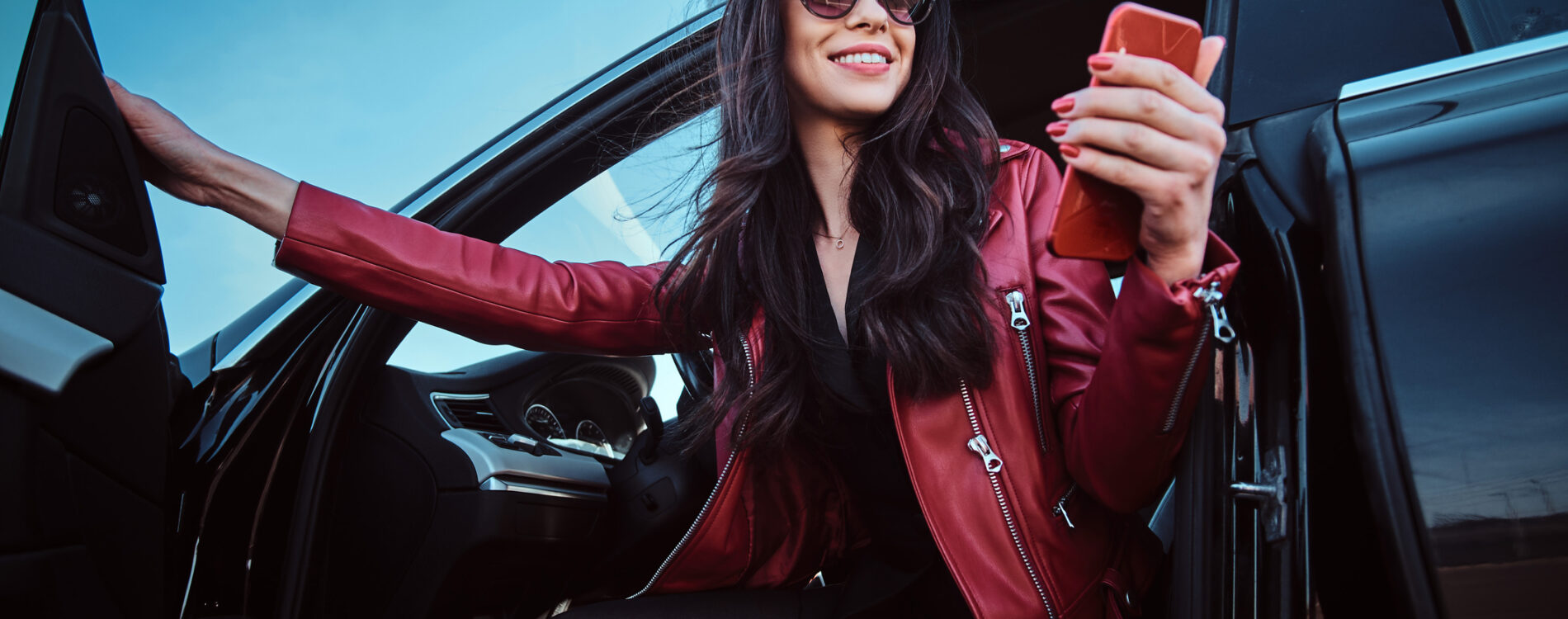 Beautiful smiling women is posing in her new car while chatting on mobile phone. She is wearing red leather jacket and sunglasses.